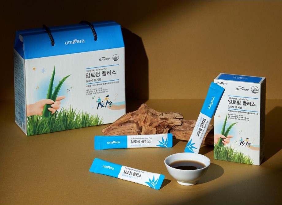 브라운 배경 앞에 놓인 알로청 플러스 선물박스와 소박스, 스틱 3포, 내용물을 담은 흰 찻잔, 고목 조각