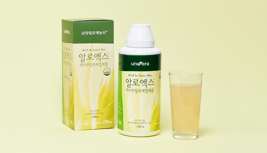 연두 벽 앞의 알로엑스 상자(남양알로에농산 · Alo-X by Active-Aloe · 알로엑스 · 액티브알로에겔제품 · 1,000 g)와 흰 뚜껑 병(univera · 알로엑스), 알로에 음료가 담긴 잔