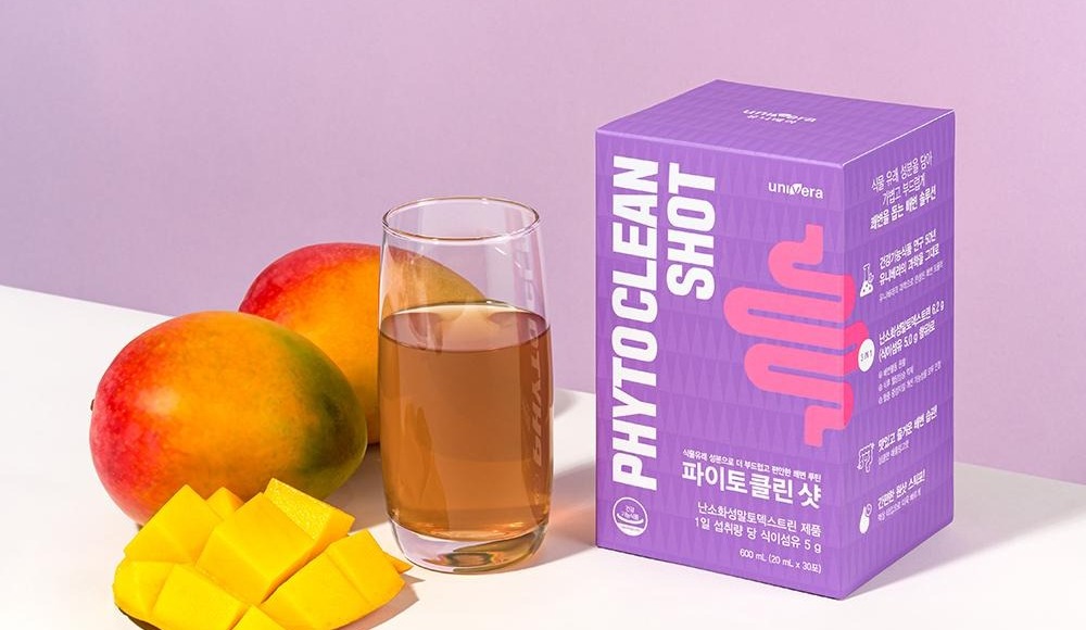 연보라 벽과 흰 테이블 위의 망고 두 개와 자른 망고, 음료가 담긴 유리컵, 파이토클린 샷 상자