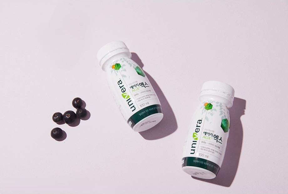 연보라 바닥에 비스듬히 놓인 에이지엑스 100 ml 병 두 개와 블랙커런트 열매