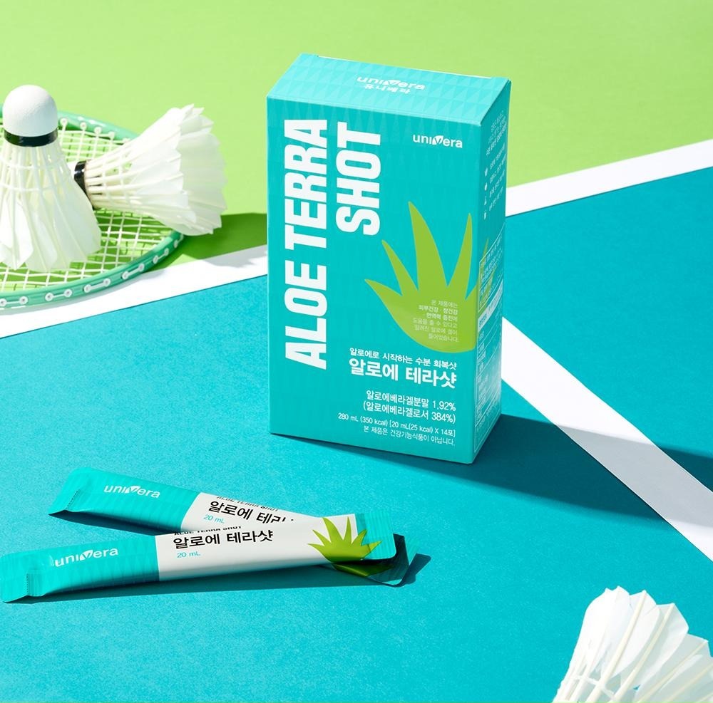 청록색 코트 위에 놓인 알로에 테라샷 상자와 액상스틱 두 포, 배드민턴 라켓과 셔틀콕
