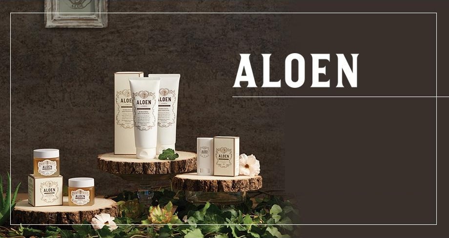 ALOEN. 짙은 갈색 벽 앞, 나무 받침과 담쟁이 위에 놓인 알로엔 제품들 — 가운데 모이스처 캡슐젤 튜브 둘과 상자, 오른쪽 선스틱과 상자, 왼쪽 나이트 밸런싱젤 단지 둘과 상자. 왼쪽 위에 낡은 흰 액자, 둘레에 가는 흰 테두리 선