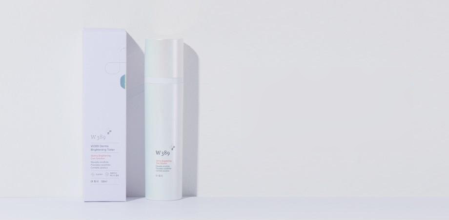 연보랏빛 회색 벽 앞 선반 위의 W389 더마 브라이트닝 토너 상자(흰 · 연보라)와 토너 병