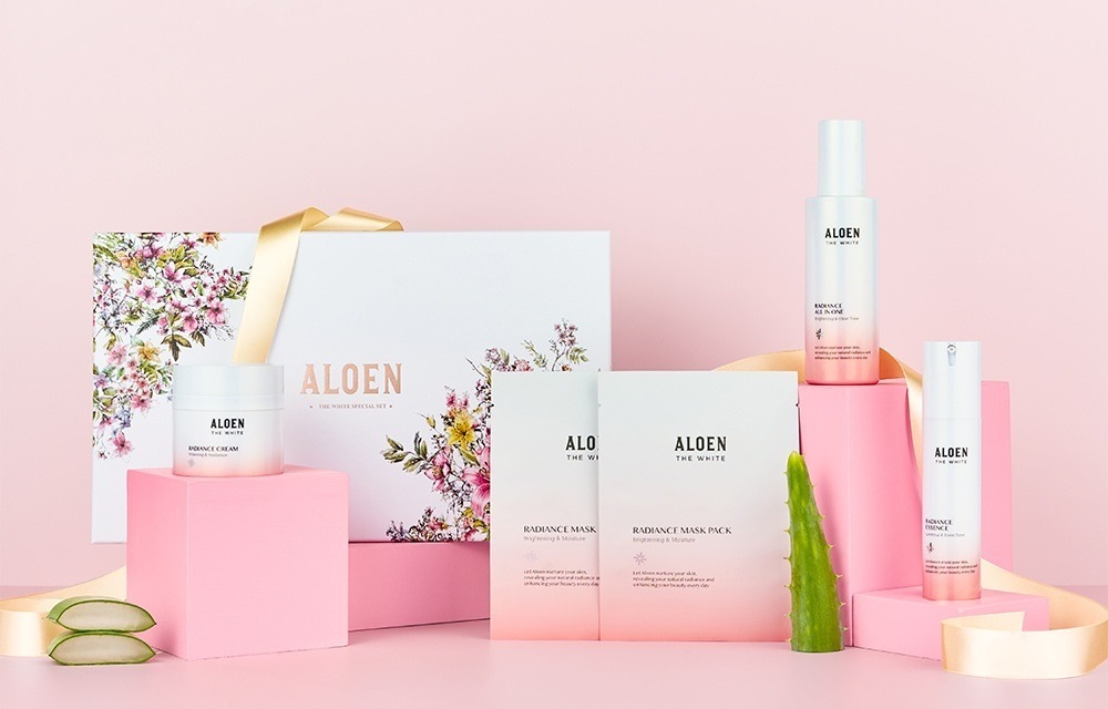 분홍 받침 위 꽃무늬 기프트 박스(ALOEN, 금빛 리본)와 알로엔 더화이트 래디언스 크림 단지 · 마스크팩 파우치 둘 · 올인원 병 · 에센스 병, 알로에 잎