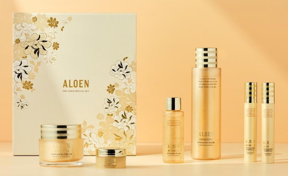 금박과 흰색 모란 무늬의 아이보리 선물 상자(ALOEN, THE GOLD SPECIAL SET) 앞에 알로엔 안티에이징 크림 50ml 와 10ml, 오른쪽에 플루이드 50ml 와 200ml, 앰플 2개가 금빛 벽과 탁자 위에 놓여 있다. 용기 글자: ALOEN, THE GOLD, ANTIAGING CREAM Elasticity & Nourished, ANTIAGING FLUID Vitality & Smoothness, ANTIAGING AMPOULE Nourished & Skin gloss