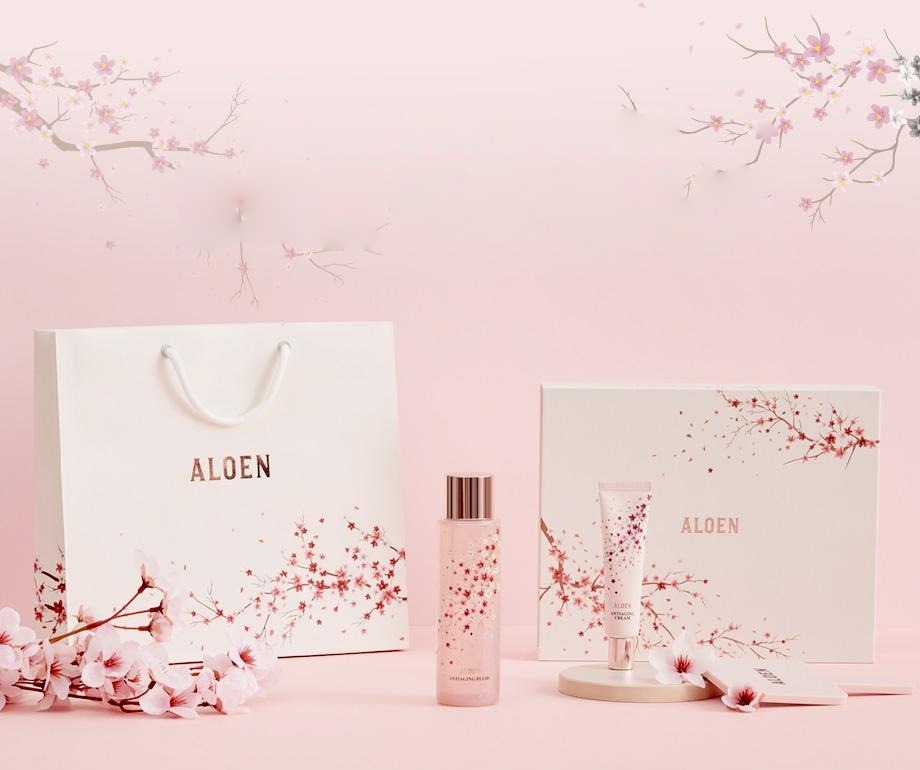 분홍 벽 앞 벚꽃 가지 아래, 벚꽃 무늬 흰 쇼핑백(ALOEN) · 벚꽃 무늬 알로엔 안티에이징 플루이드 병 · 안티에이징 크림 튜브 · 벚꽃 무늬 선물 상자(ALOEN) · 둥근 받침과 손거울 · 벚꽃 송이. 용기 글자: ALOEN, ANTIAGING FLUID, ANTIAGING CREAM
