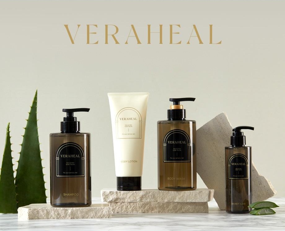 회베이지 벽 위쪽의 금색 VERAHEAL 워드마크. 아래는 알로에 잎과 돌 받침 위에 선 베라힐 라인 넷 — SHAMPOO(펌프 병) · BODY LOTION(흰 튜브) · BODY WASH(펌프 병) · SECRET CARE(작은 펌프 병)